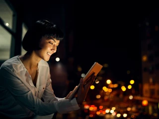 picture of a smiling lady looking at her tablet sat on a balcony in a city at night